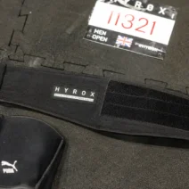 Black nylon lifting belt for HYROX laid on rubber race floor tiles next to a race bib and knee sleevesVous avez dit : Ca correspond bien?