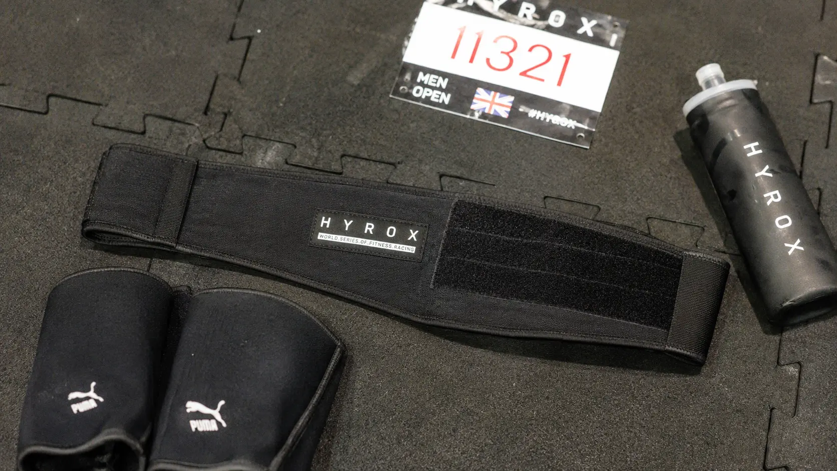 Black nylon lifting belt for HYROX laid on rubber race floor tiles next to a race bib and knee sleevesVous avez dit : Ca correspond bien?