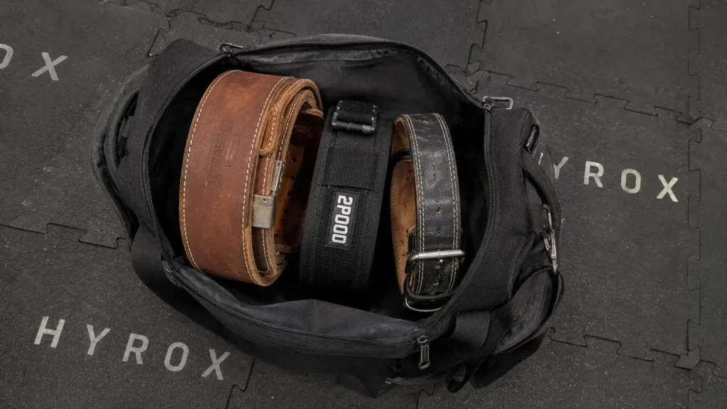 Open gym bag with multiple weightlifting belts showing the difference between a powerlifting belt and a nylon lifting belt for HYROX