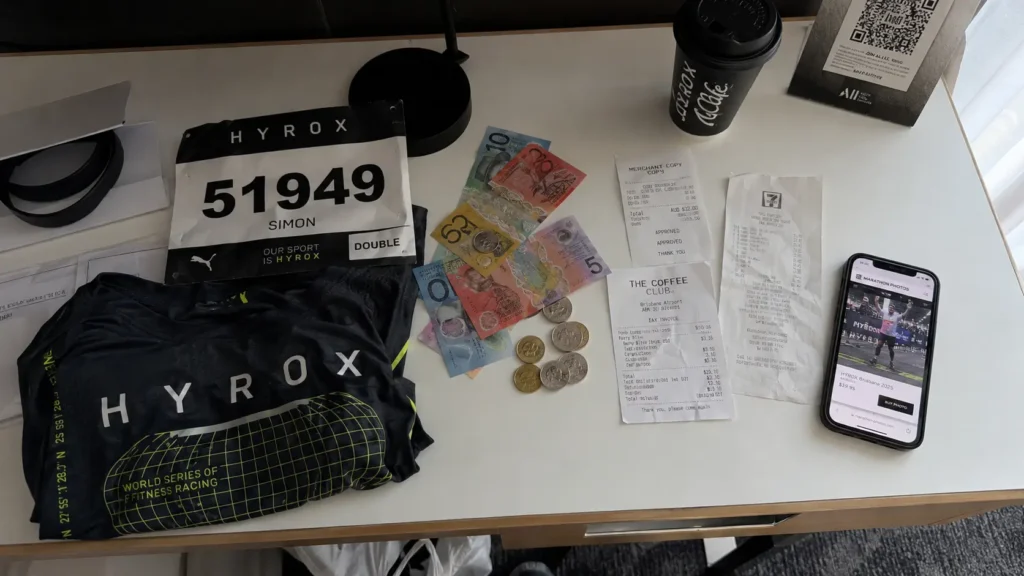 Overhead flat-lay of Australian cash, race bib, receipts and HYROX merchandise on hotel desk — the hidden costs that push HYROX cost in Australia over budget